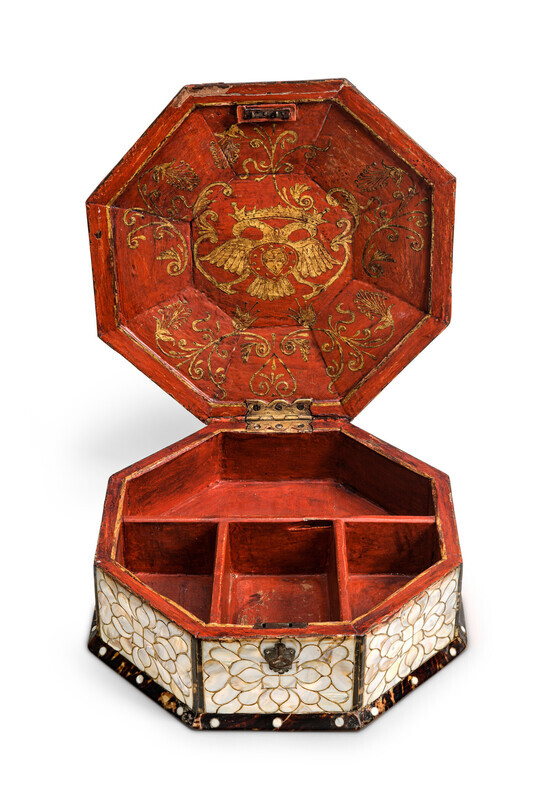 Open octogonal box with ornate mother-of-pearl inlay in an abstract floral pattern framed by tortoiseshell, displaying red painted interior with gold detialing of a coat of arms. Photographed on a white background. 