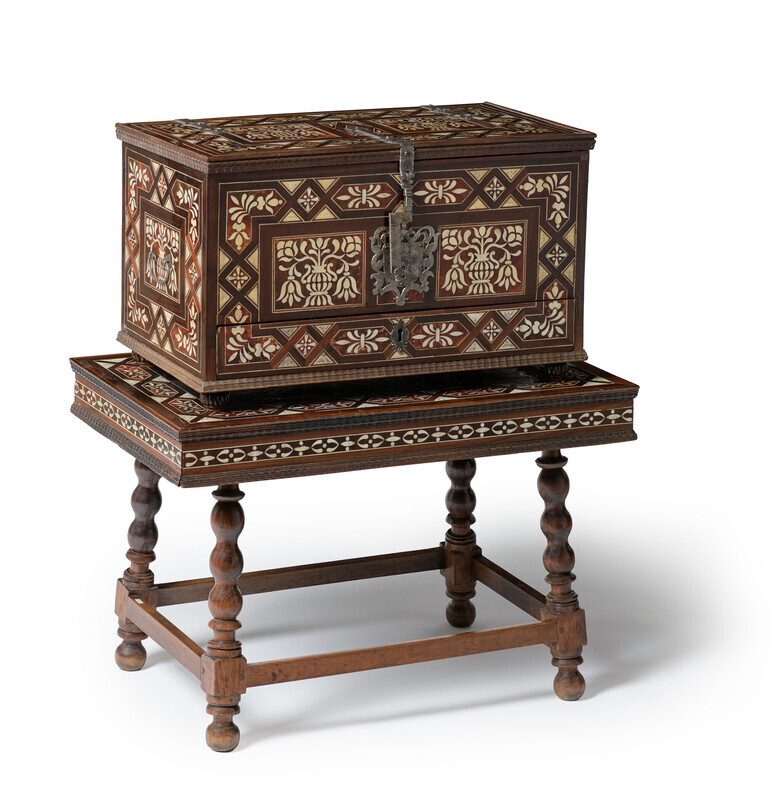 Ornate, rectangular chest in wood on a matching four-legged stand with tortoiseshell, mother-of-pearl, and ivory inlay in floral and geometric pattersn and iron fittings with the top and drawers closed. The objects are photographed on a white background.