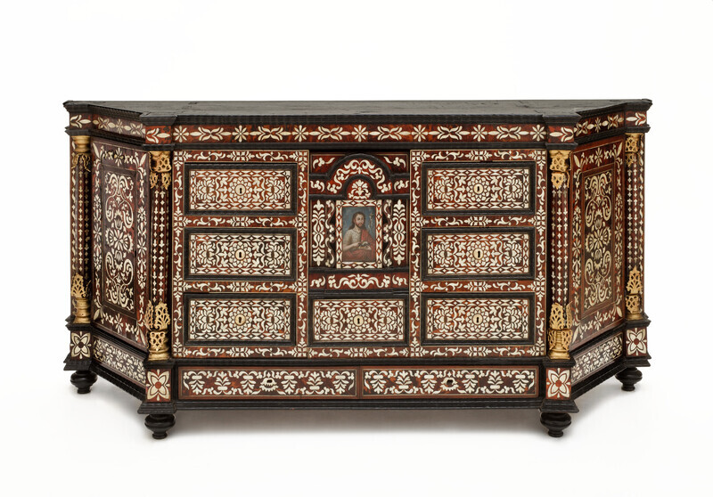 Low, rhombus shaped wooden cabinet with tortoiseshell and mother-of-pearl inlay in abstract floral pattern. In center of the front panel is a painting of a bearded man with long brown hair wearing a gray hair shirt and a red tunic. The man has a halo, carries a crucifix, a bible, and a sheep figurine. The object is photographed on a white background. 