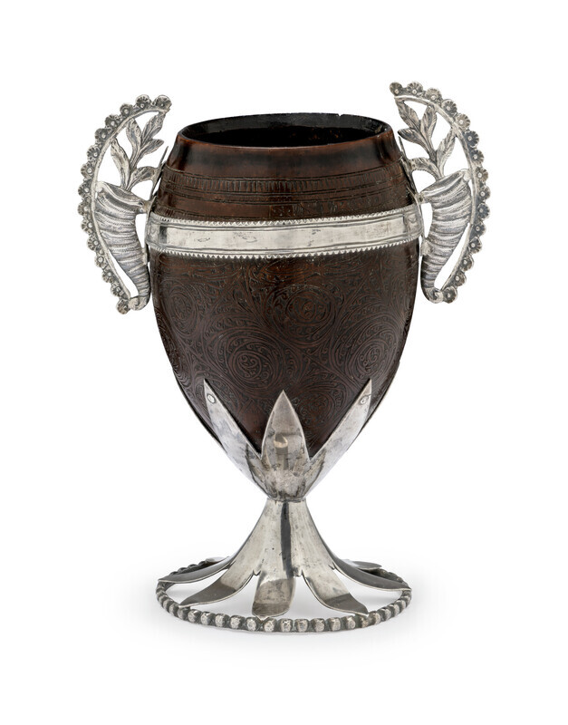 Ornate cup made of a polished coconut with encised floral decoration, mounted with curling silver handles on either side made of cornucopieas, a silver stem, and a circular silver base. Photographed on a white background. 