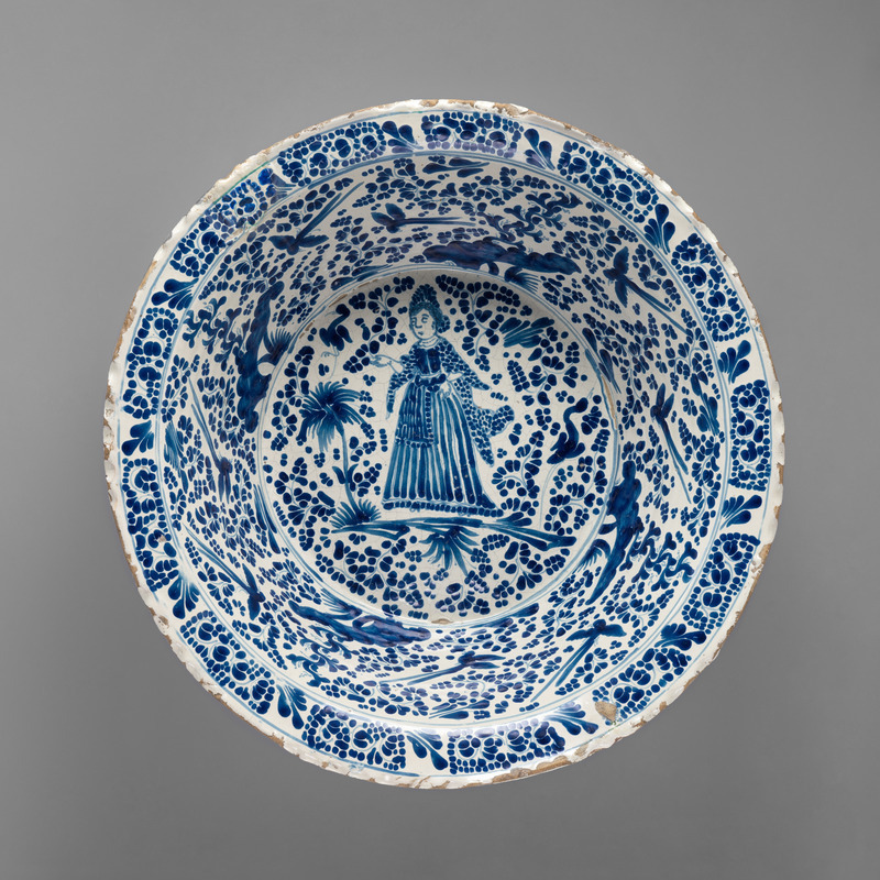 Tin-glazed ceramic basin with an extended lip seen from above and photographed on a neutral gray background. Decorated with blue-on-white glaze with birds and flowers on the sides of the interior and a woman on the bottom of the base.