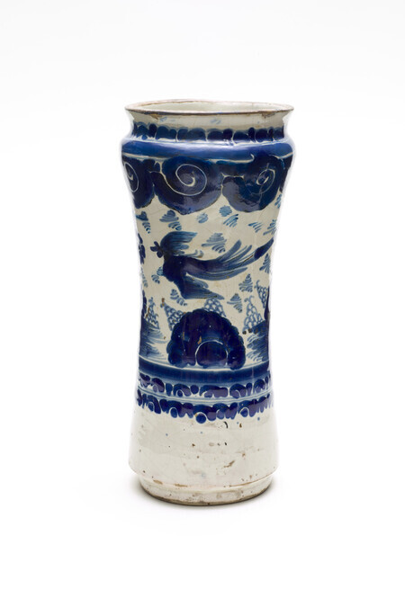 Tin-glazed ceramic jar that flares at the top and the bottom. Decorated in blue-on-white glaze with a bird flying over a landscape with clouds in the sky. Photographed on a white background.