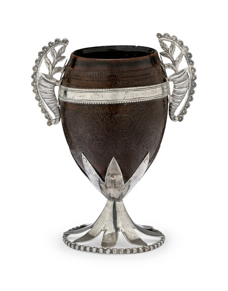 Ornate cup made of a polished coconut with encised floral decoration, mounted with curling silver handles on either side made of cornucopieas, a silver stem, and a circular silver base. Photographed on a white background. 